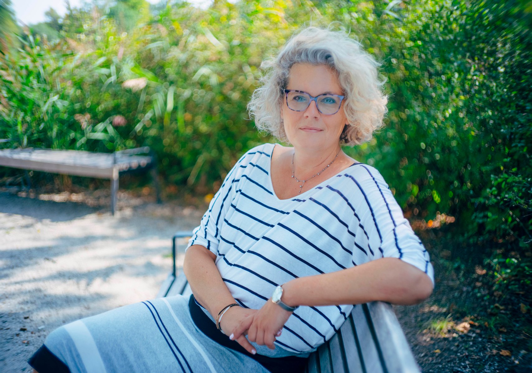 A woman with light, curly hair and glasses sits on a park bench. - conversation graphic