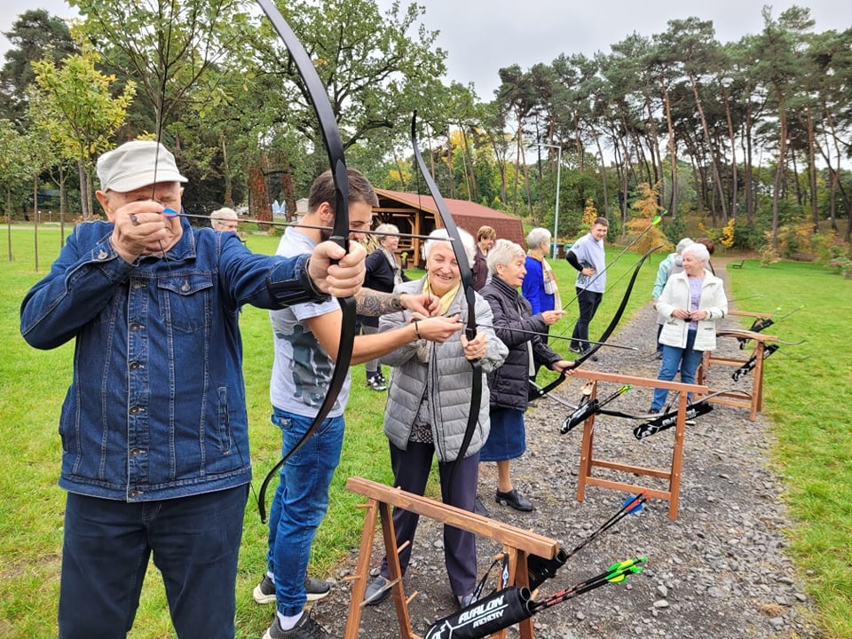 Teren zielony, w oddali drzewa. Mężczyzna strzela z kuszy. Obok młodszy mężczyzna pokazuje kobiecie, jak strzelać z kuszy. Kilka osób przygląda się temu. - grafika rozmowy