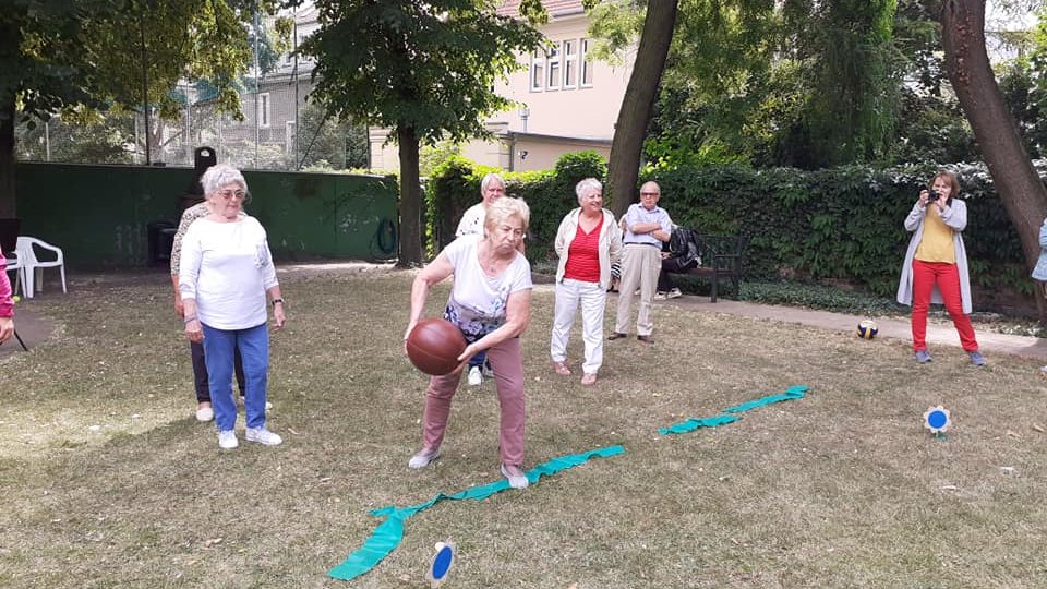 Ogród. Mężczyzna i kilka kobiet przyglądają się kobiecie, która rzuca piłkę.