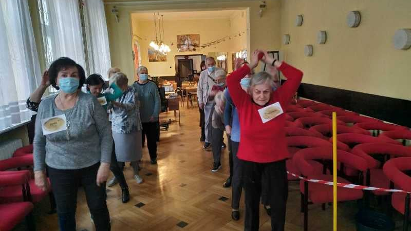 Pomieszczenie. Seniorzy stoją w dwóch rzędach jeden za drugim.