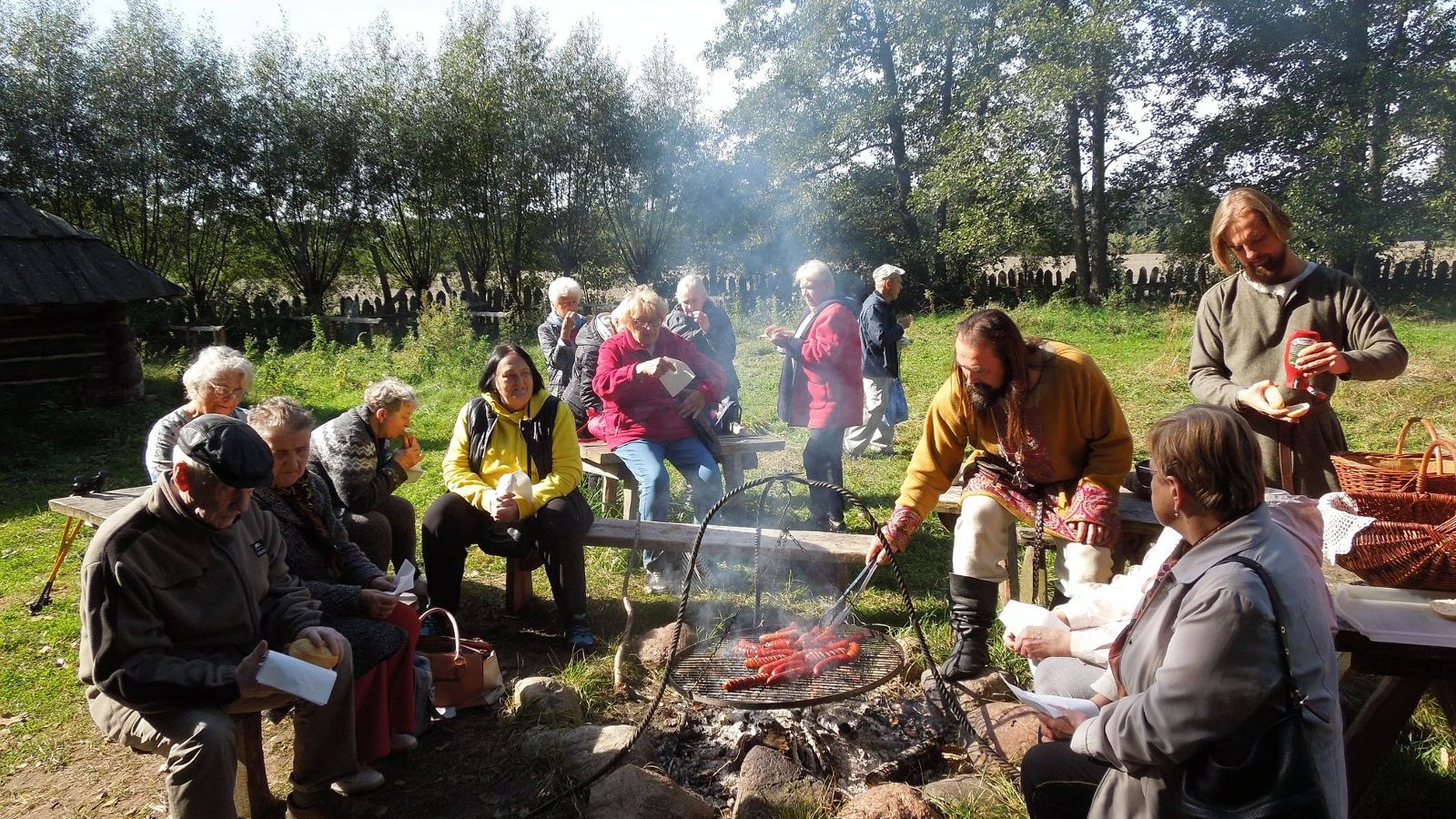 Plener, w dali drzewa. Ludzie siedzą wokół ogniska, na ruszcie pieką się kiełbaski.