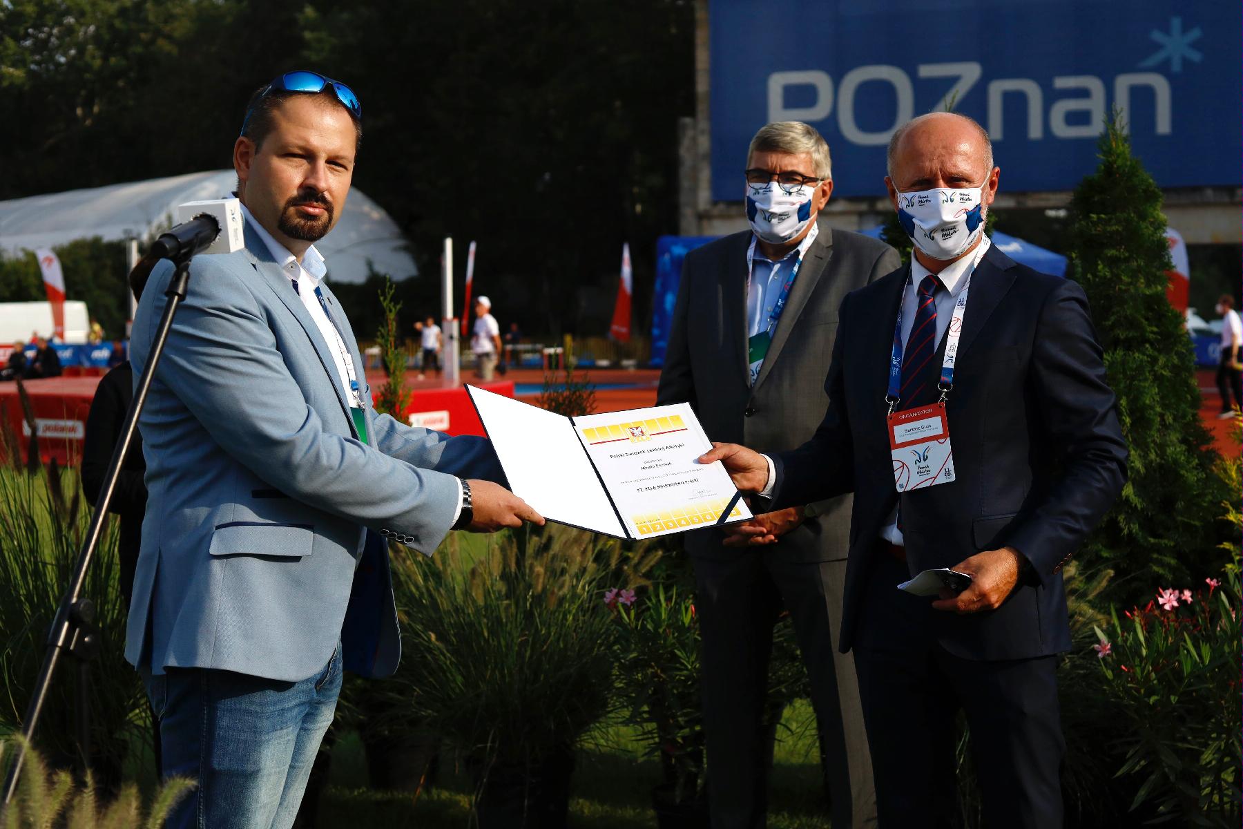 Przedstawiciel PZLA wręcza Bartoszowi Gussowi certyfikat przyznania Poznaniowi organizację Mistrzostw Polski w lekkoatletyce w 2021. Na drugim planie Grzegorz Ganowicz, przewodniczący Rady Miasta - grafika rozmowy