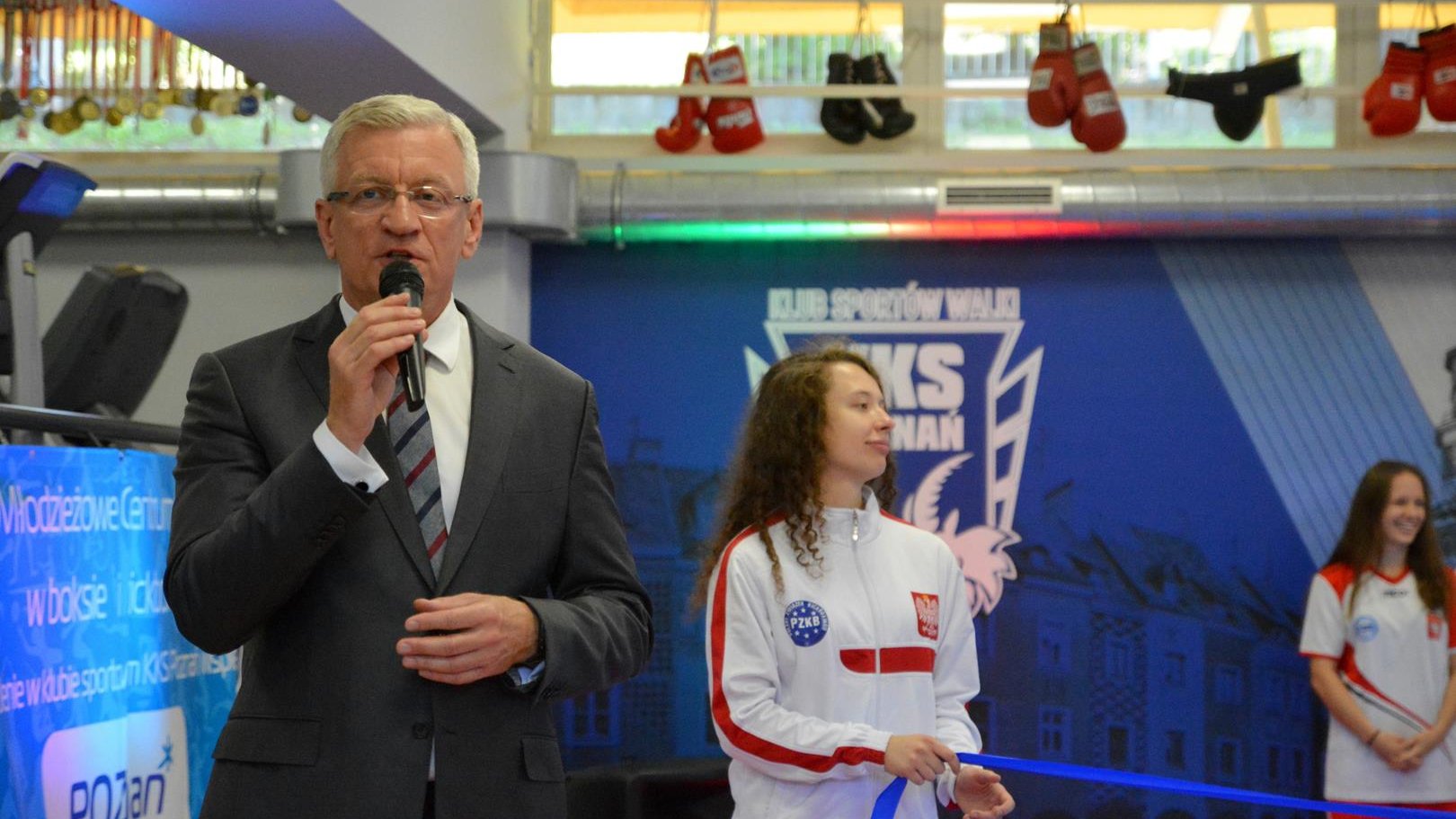 Jacek Jaśkowiak, prezydent Poznania, przy mikrofonie. W tle zawodniczka KKS Poznań, trzymająca granatową wstęgę, która wkrótce ma zostać przecięta