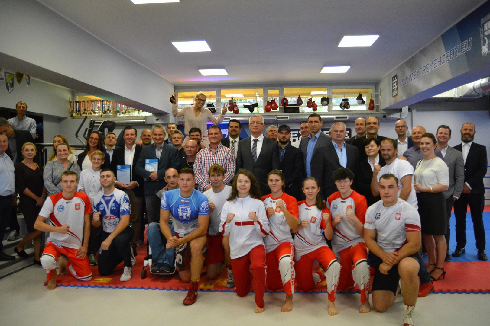 Duża grupa ludzi pozuje do zdjęcia w nowej sali. W pierwszym rzędzie klęczą zawodnicy KKS Poznań w pozach sportowych, w centrum Jacek Jaśkowiak, Radosław Laskowski i Jędrzej Solarski - grafika rozmowy