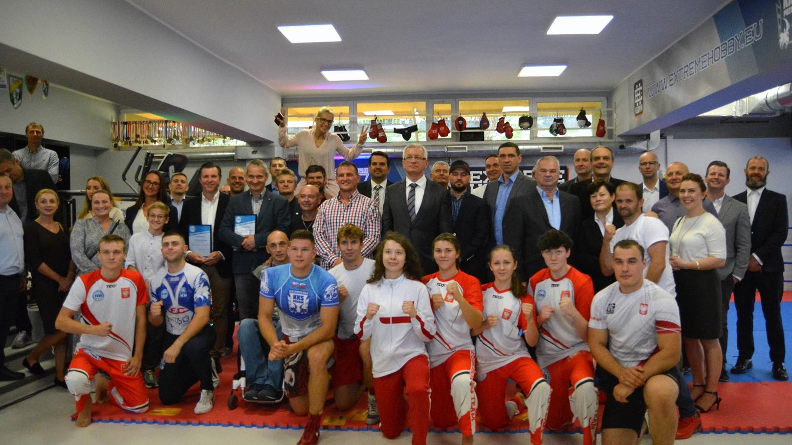 Duża grupa ludzi pozuje do zdjęcia w nowej sali. W pierwszym rzędzie klęczą zawodnicy KKS Poznań w pozach sportowych, w centrum Jacek Jaśkowiak, Radosław Laskowski i Jędrzej Solarski