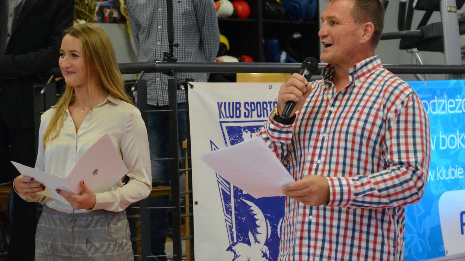 Na zdjęciu przy mikrofonie Radosław Laskowski, prezes Klubu sportów Walki, uśmiechnięty, z kartką w dłoni. Obok po lewej młoda kobieta o jasnych włosach, z teczką w ręku