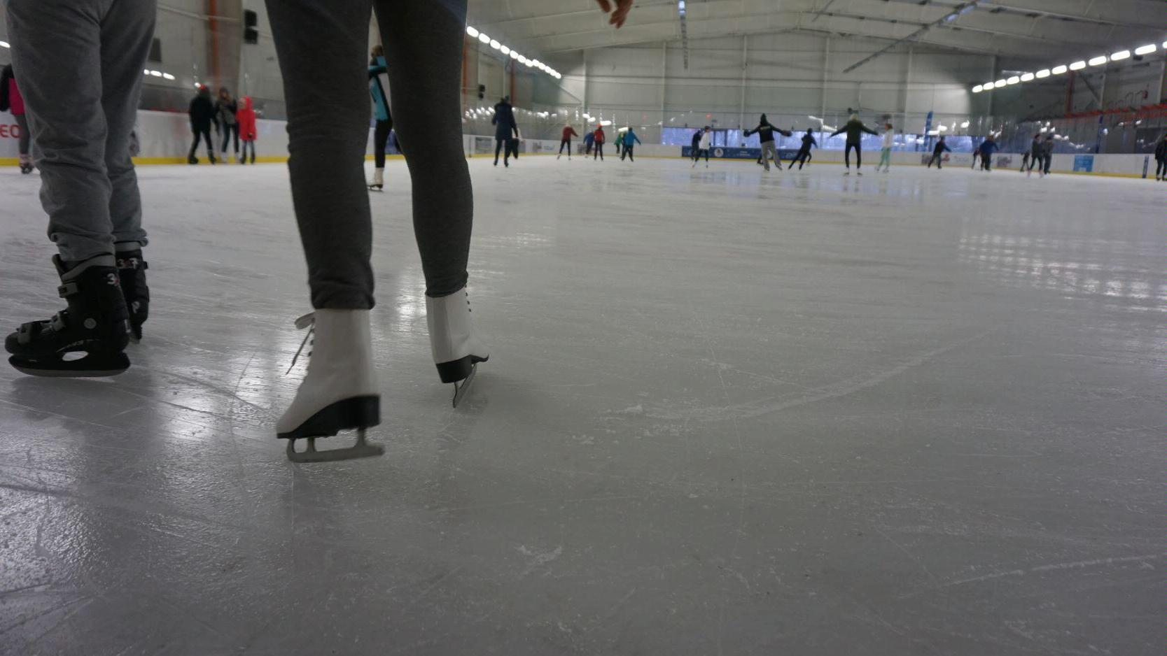 Zdjęcie przedstawia lodowisko. Na pierwszym planie widać nogi, na których znajdują się łyżwy. W tle znajdują się osoby, które jeżdżą na łyżwach. - grafika rozmowy
