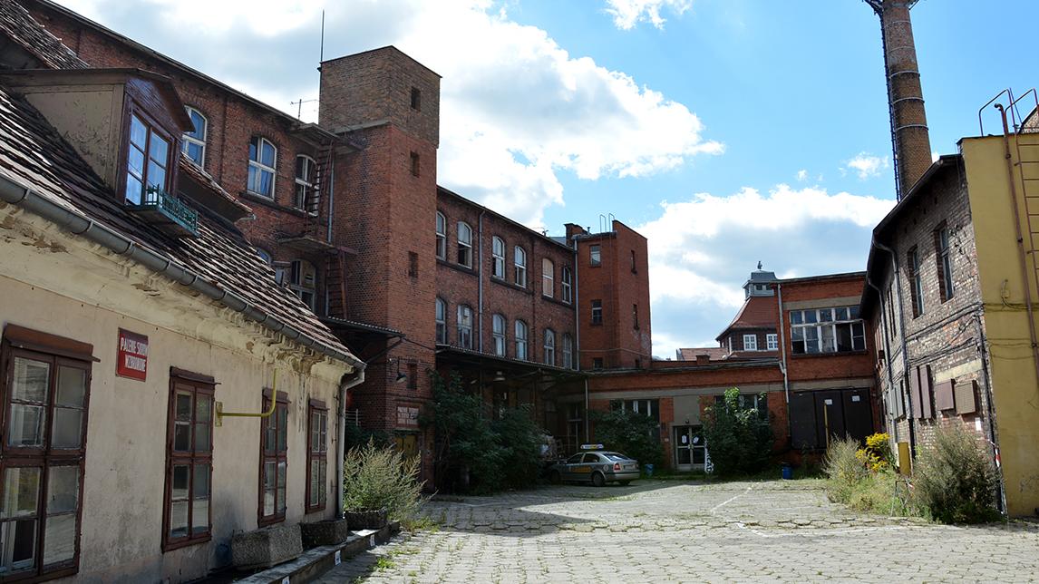 Zdjęcie przedstawia tereny Starej Papierni: po lewej stronie niski tynkowany budynek, w centrum budowle z cegły - grafika rozmowy