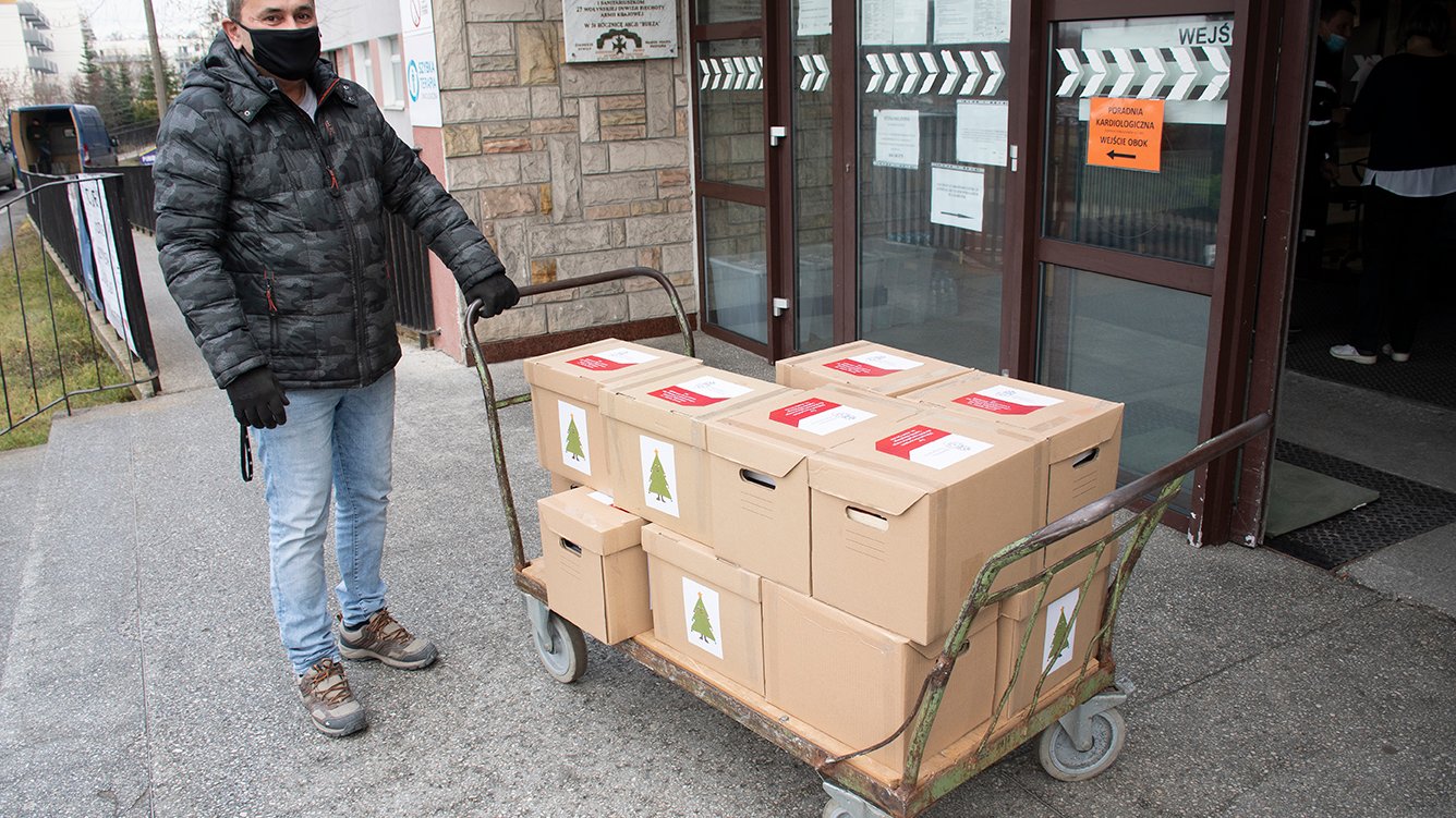 Zdjęcie przedstawia człowieka pchającego wózek, na którym znajdują się kartony z książkami. Człowiek ten stoi przed wejściem do szpitala.