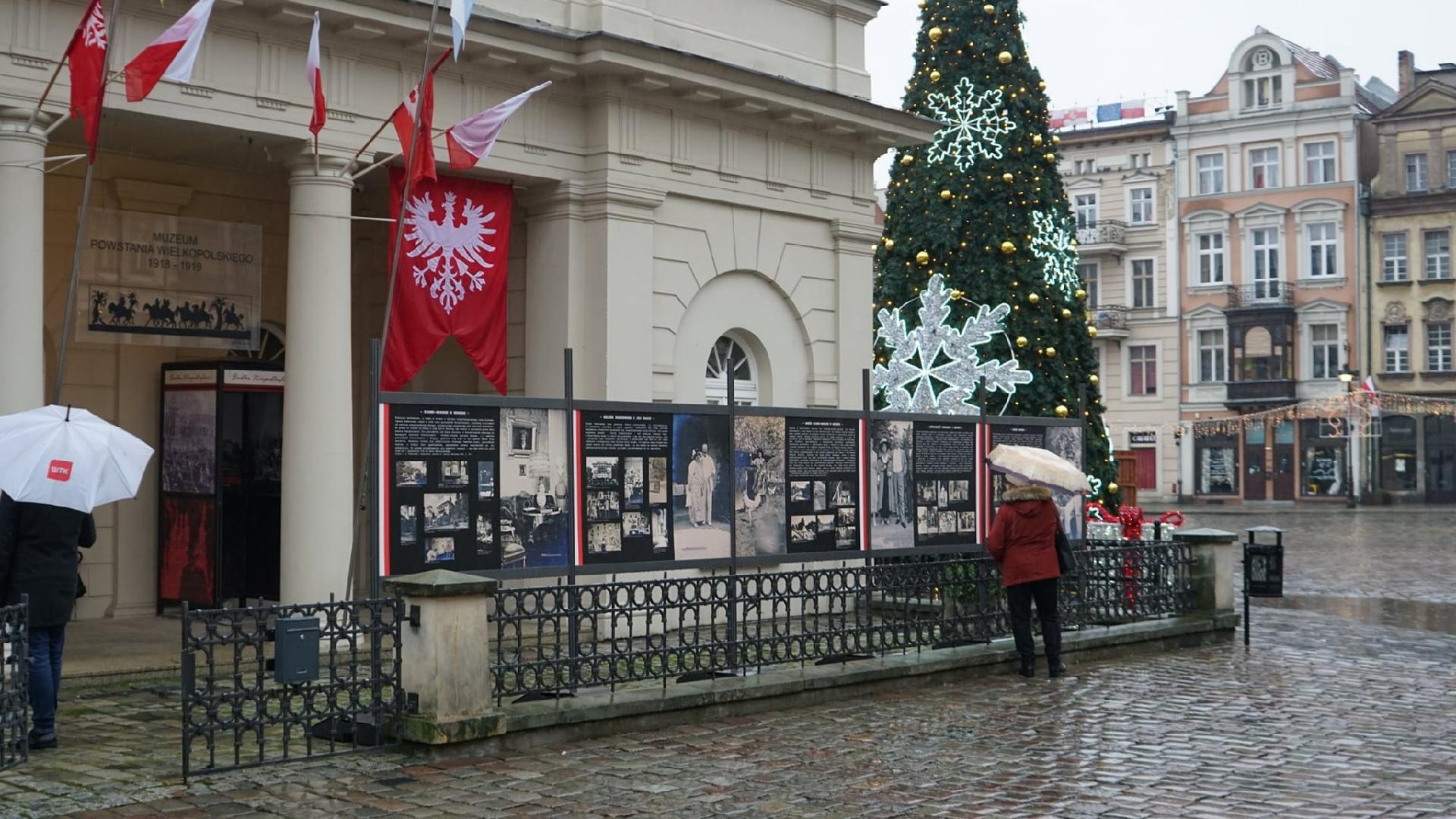 Stary Rynek: na pierwszym planie widzimy budynek muzeum, przed nim znajdują się tablice ze zdjęciami Paderewskiego. Wystawę oglądają przechodnie z parasolami. Widać też choinkę. - grafika rozmowy