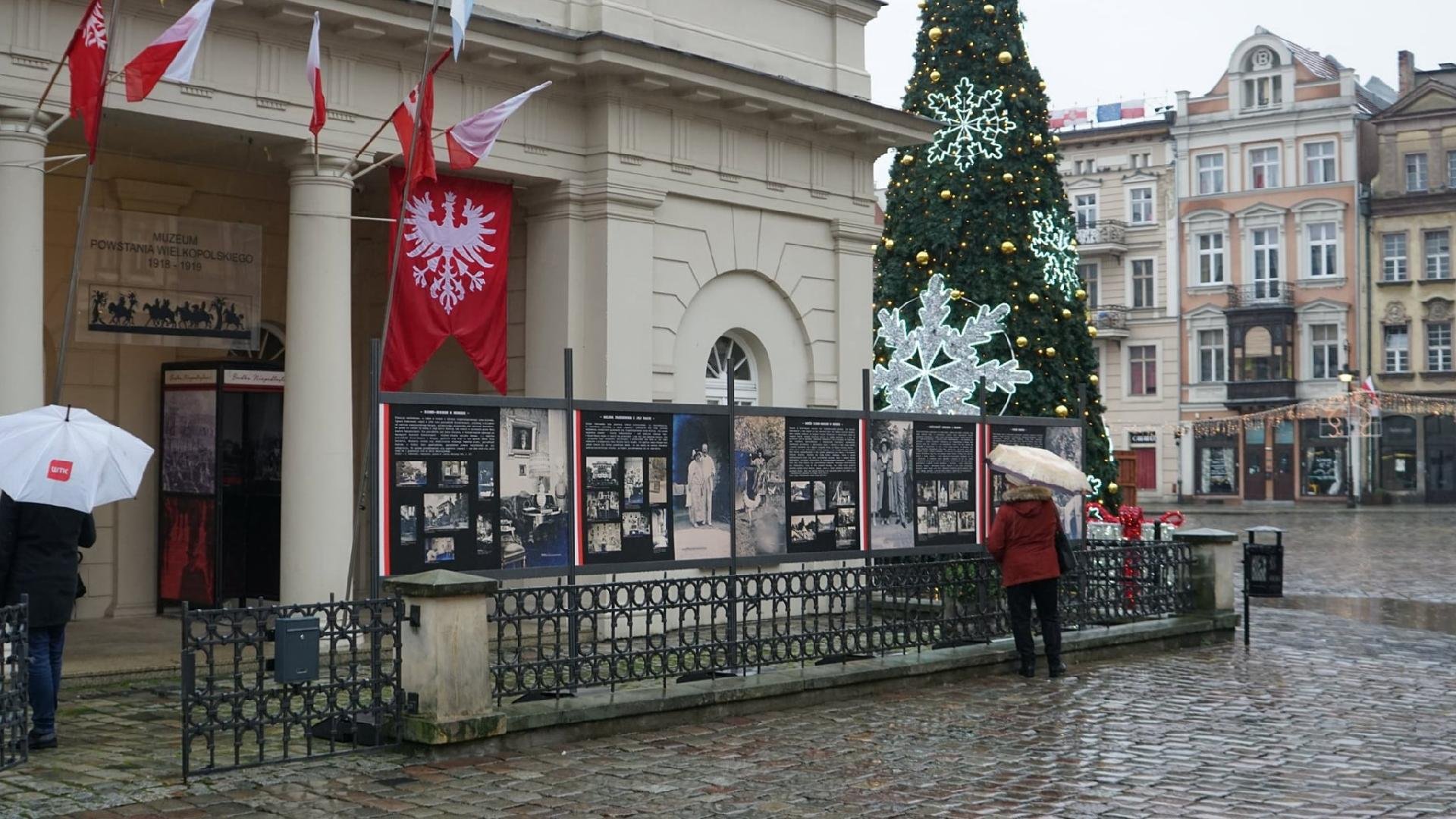 Stary Rynek: na pierwszym planie widzimy budynek muzeum, przed nim znajdują się tablice ze zdjęciami Paderewskiego. Wystawę oglądają przechodnie z parasolami. Widać też choinkę.