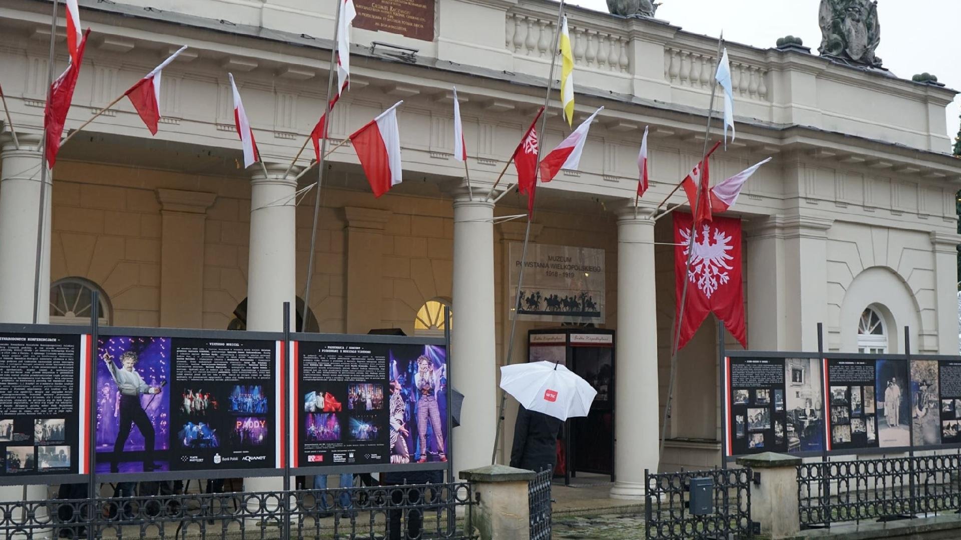 Stary Rynek: na pierwszym planie widzimy budynek muzeum, przed nim znajdują się tablice ze zdjęciami Paderewskiego. Wystawę oglądają przechodnie z parasolami.