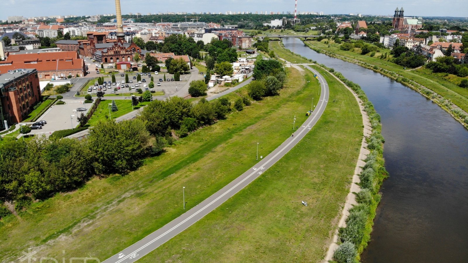 Zdjęcie przedstawia Wartostradę. Widać na nim drogę rowerową prowadzącą przez tereny zieleni. W sąsiedztwie znajdują się się zabudowania i rzeka.