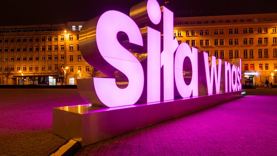 Zdjęcie przedstawia różowy, podświetlony neon z napisem "Siła w nas". Znajduje się on na placu Wolności. Zdjęcie zostało zrobione wieczorem.