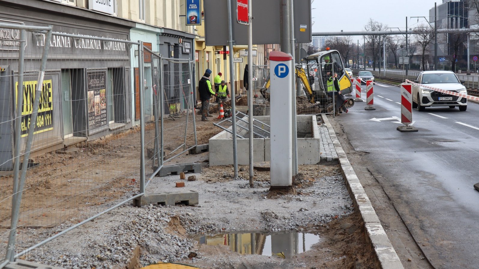 Pracownicy budowlani układają nawierzchnię chodnika. Na zdjęciu widać też jezdnię i samochody.