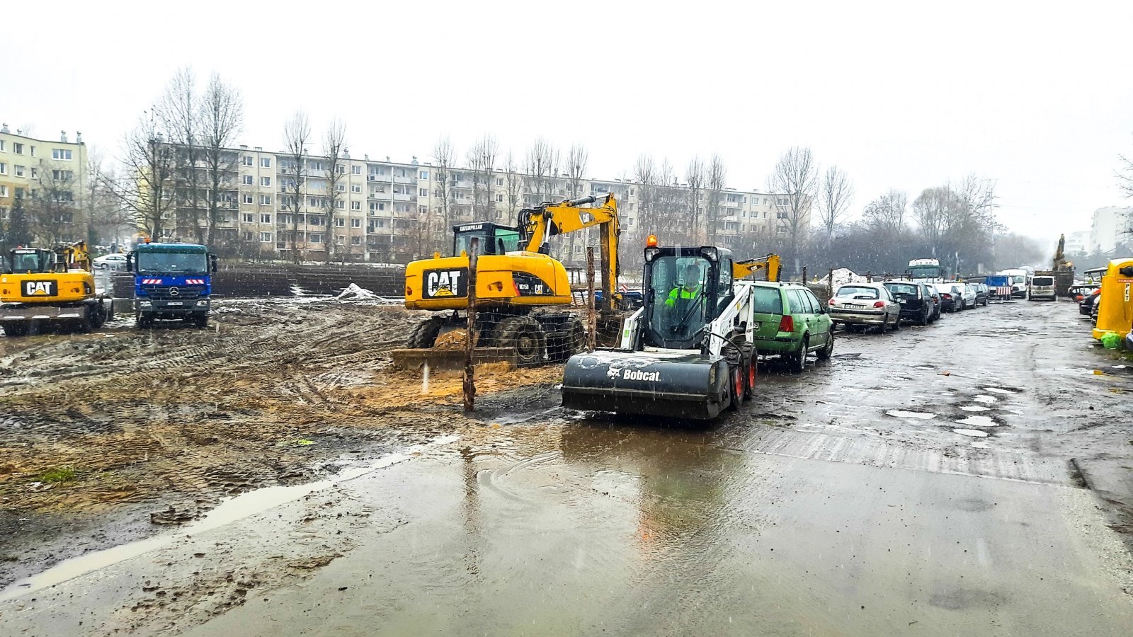 Maszyny budowalne, robotnicy i wykopy. W tle bloki mieszkalne