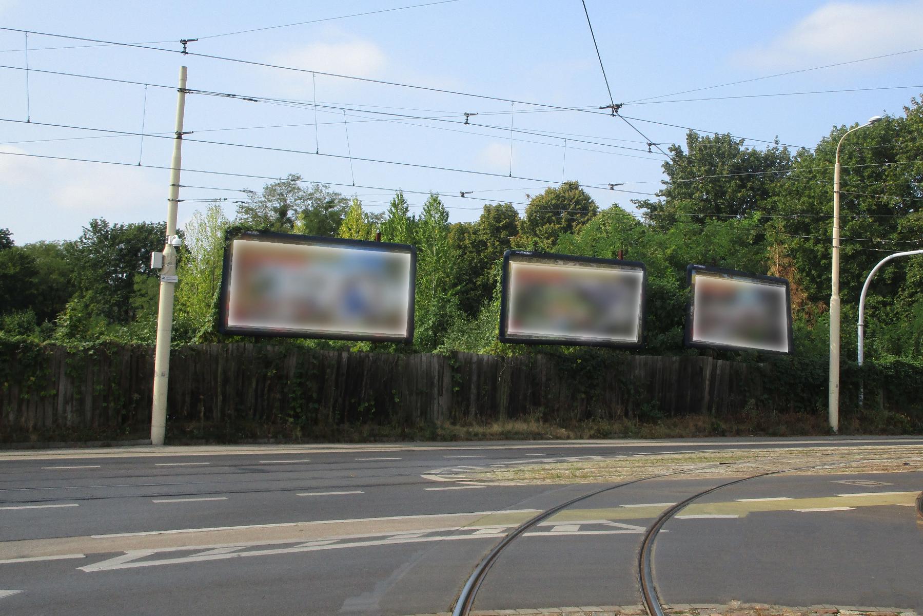 Na pierwszym planie droga, za nią płot, ponad nim wielkoformatowe banery reklamowe. Powyżej sieć trakcyjna. - grafika rozmowy