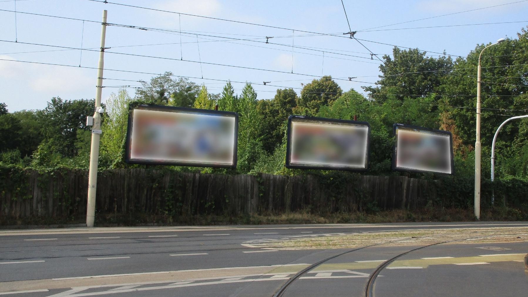 Na pierwszym planie droga, za nią płot, ponad nim wielkoformatowe banery reklamowe. Powyżej sieć trakcyjna.