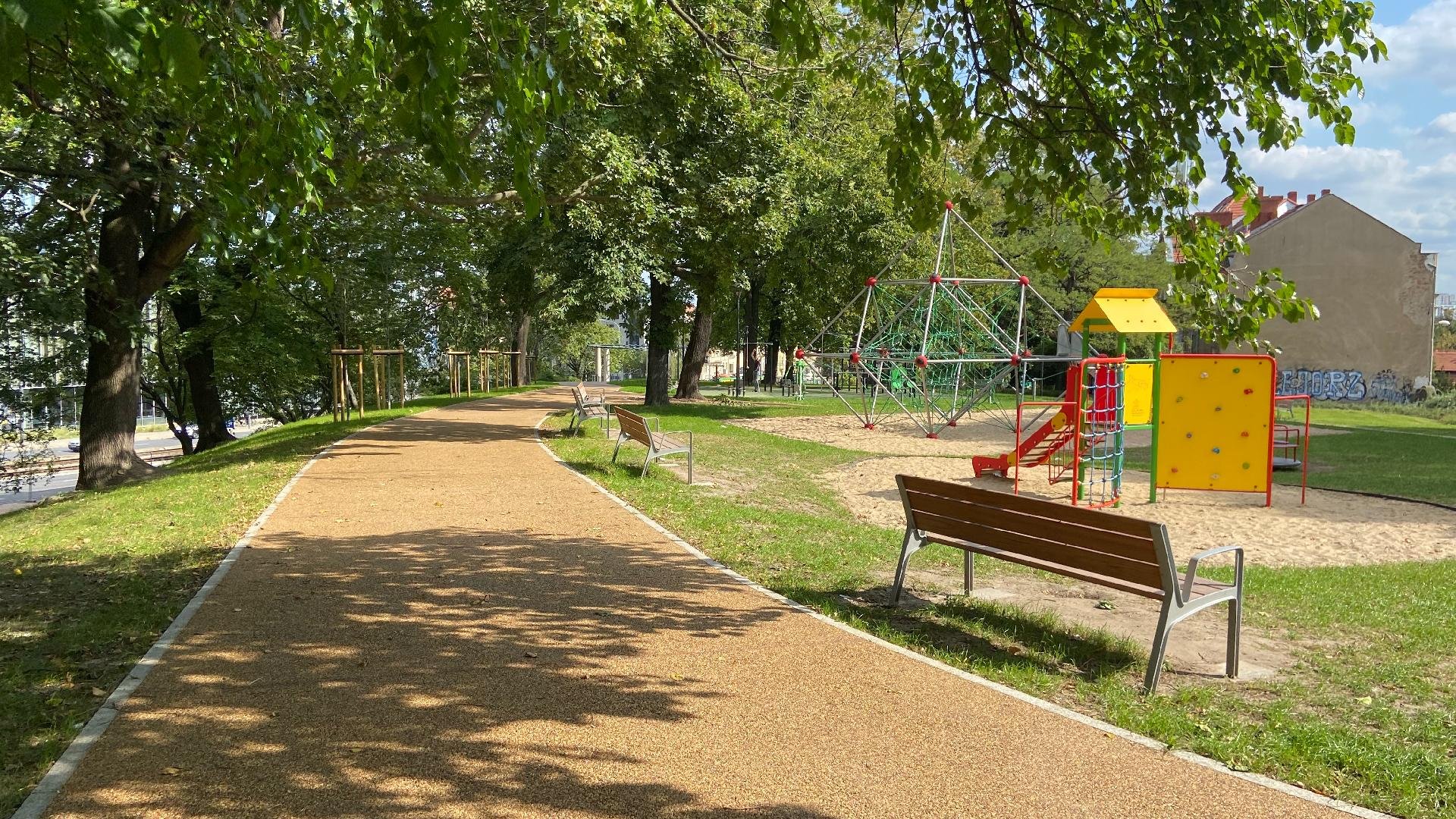 Galeria zdjęć przedstawia park Drwęskich. Na zdjęciu widać zieleń (trawę i drzewa), alejki spacerowe oraz plac zabaw, otoczony ławkami.