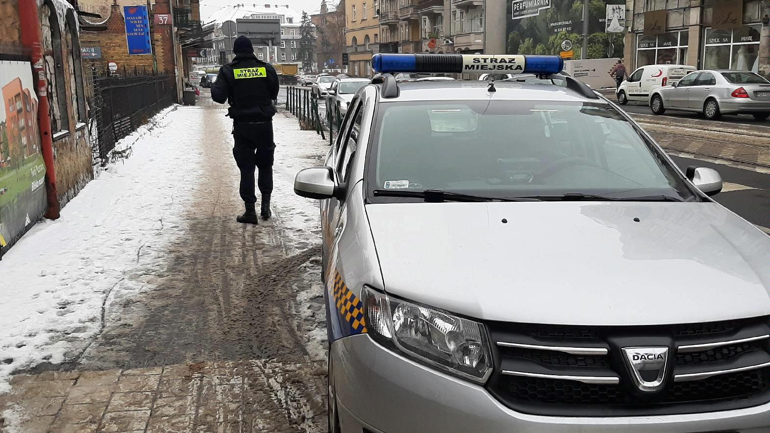 Na pierwszym planie znajduje się radiowóz straży miejskiej, stojący na poboczu przy jezdni. Na chodniku tyłem stoi strażnik miejski. W tle widać jadące po ulicy samochody oraz stojące przy drodze budynki. - grafika rozmowy