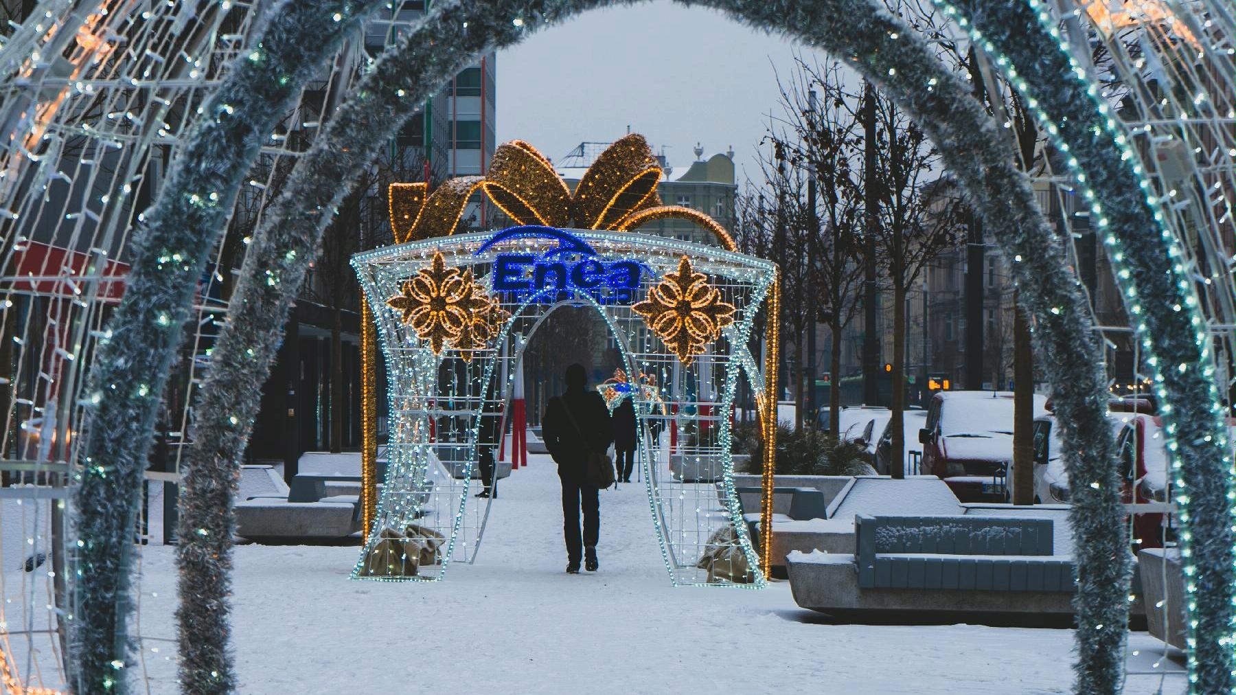 Zdjęcie przedstawia ozdoby i iluminacje świąteczne na ul. Św. Marcin. Na pierwszym planie widać świecącą dużą ozdobę w kształcie prezentu z napisem Enea, pod którą przechodzi mężczyzna w czarnym płaszczu. - grafika rozmowy