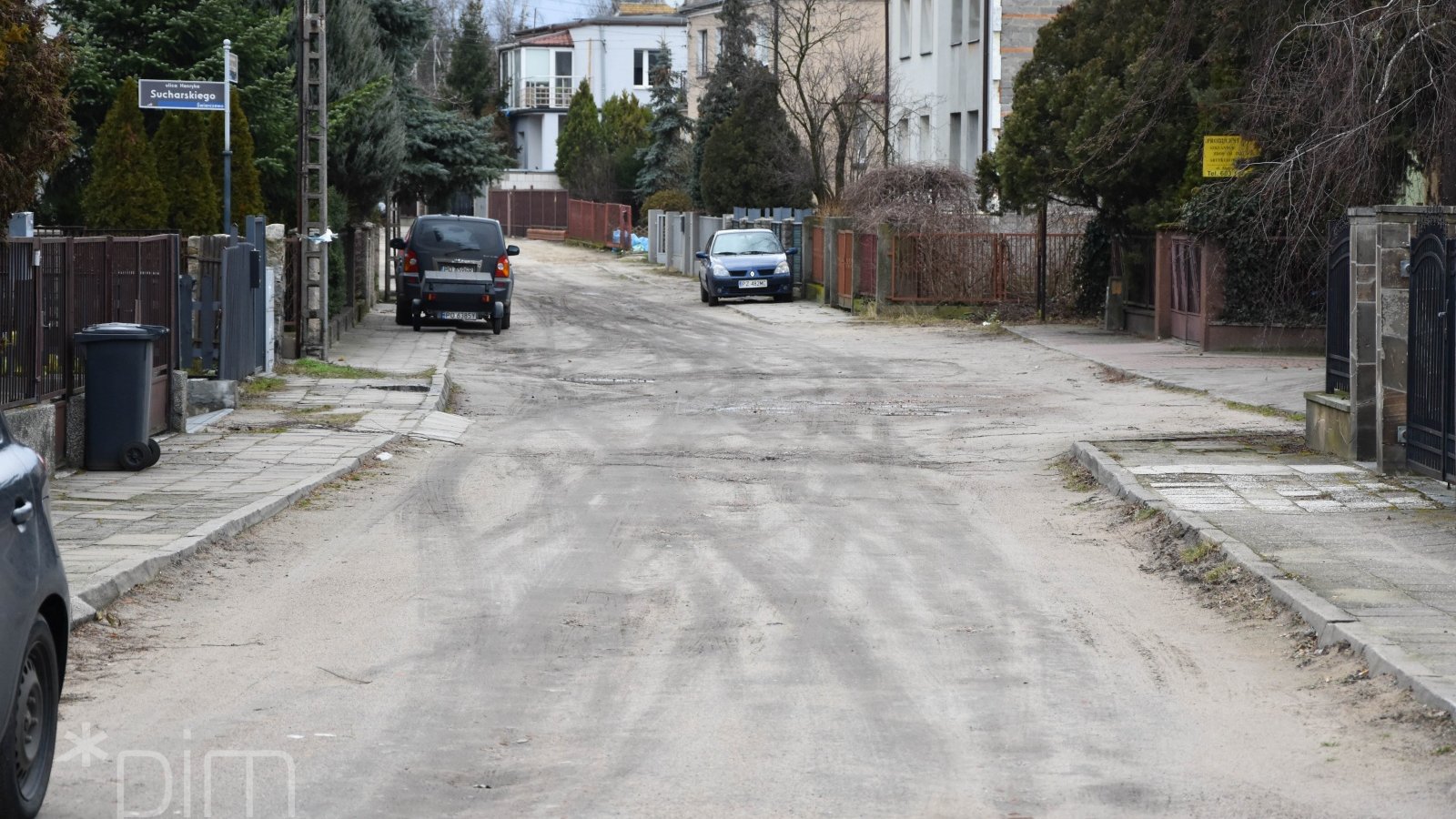 Na zdjęciu piaszczysta ulica, po jej obu stronach domy jednorodzinne, w tle zaparkowane samochody