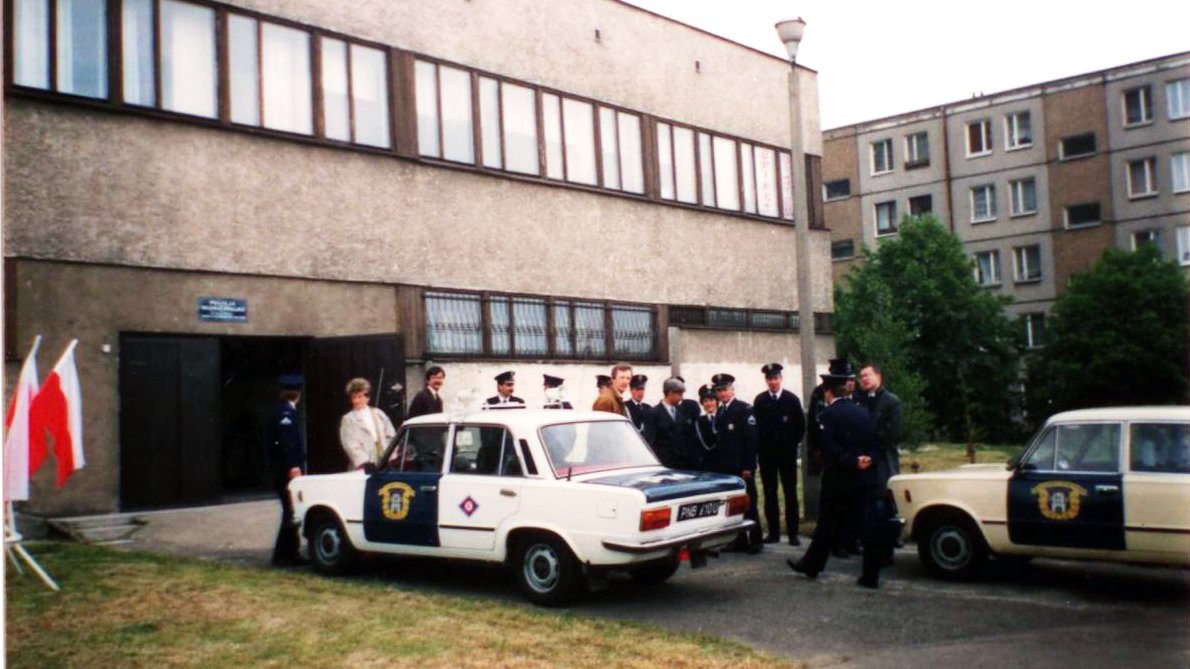 Zdjęcie przedstawia grupę osób stojących obok dwóch samochodów Straży Miejskiej. Obok znajduje się komenda oraz blok.