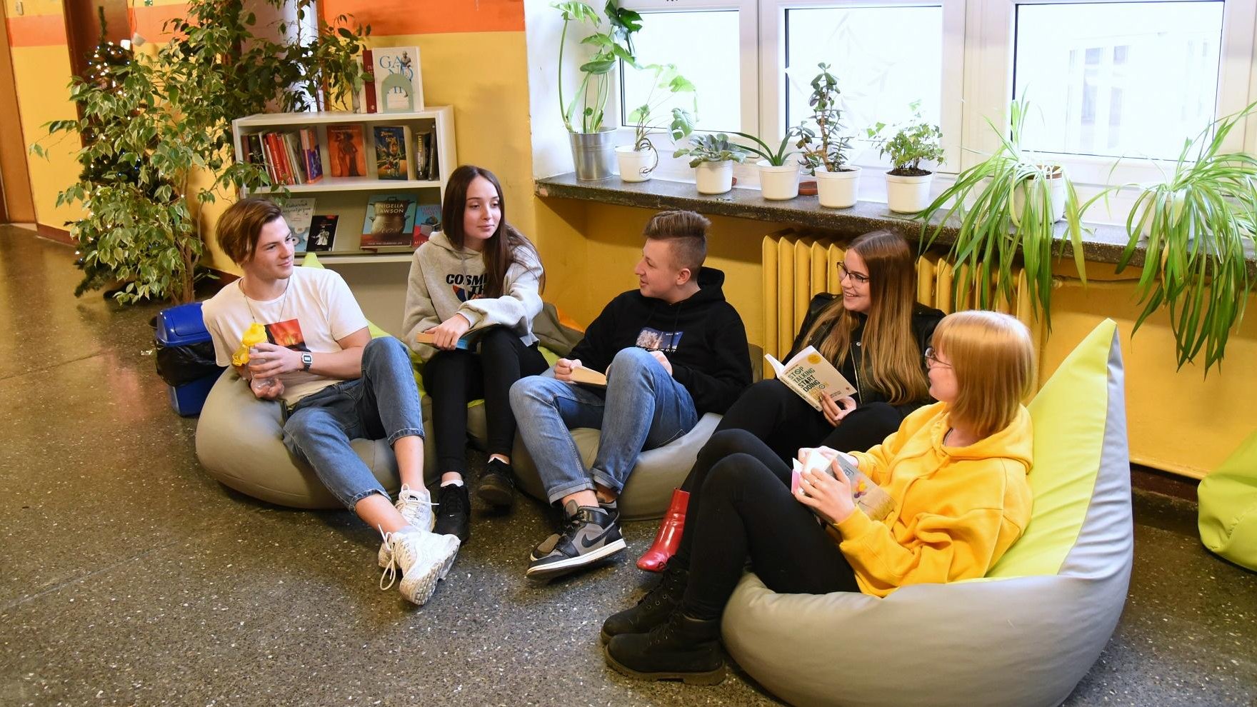 Strefa relaksu w szkole. Zdjęcie przedstawia uczniów siedzących na pufach na szkolnym korytarzu. Za ich plecami widać biblioteczkę z książkami oraz doniczki z kwiatami, stojące na parapecie.