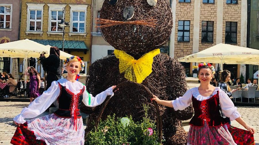 Zdjęcie przedstawia dwie kobiety ubrane na ludowo. Stoją one obok ozdoby - ogromnego zająca, w ręku trzymają duży koszyk z roślinami. Zdjęcie zostało zrobione na poznańskim Starym Rynku.