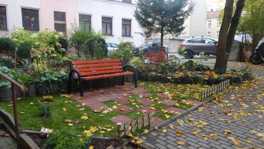 Podwórko po zakończeniu akcji. Na pierwszym planie zieleń i ławka, w tle budynek i samochody.