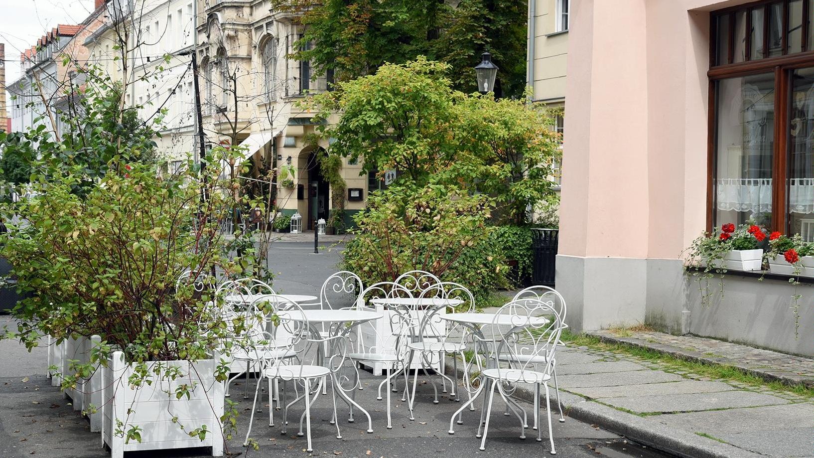 Zdjęcie ogródka gastronomicznego, na pierwszym planie białe krzesła, stolik, wokół nich rośliny