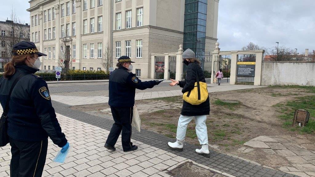 Na zdjęciu dwie strażniczki, jedna z nich wręcza maski przechodzącej ulicą kobiecie z zółtą torbą