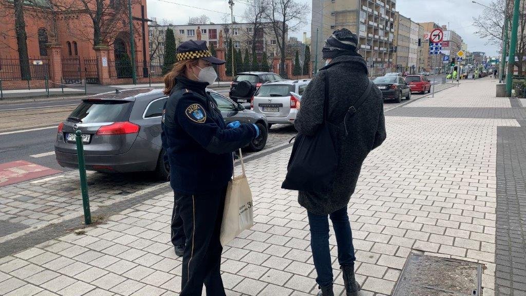 Na zdjęciu strażniczka miejska, rozdająca na ulicy maseczki - podaje je przechodniowi w szarym płaszczu