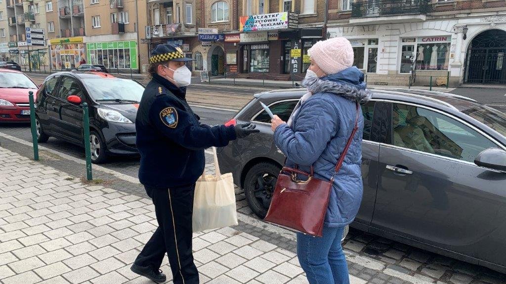 Na zdjęciu strażnik miejski podaje maseczkę przechodzącej ulicą kobiecie w niebieskiej kurtce