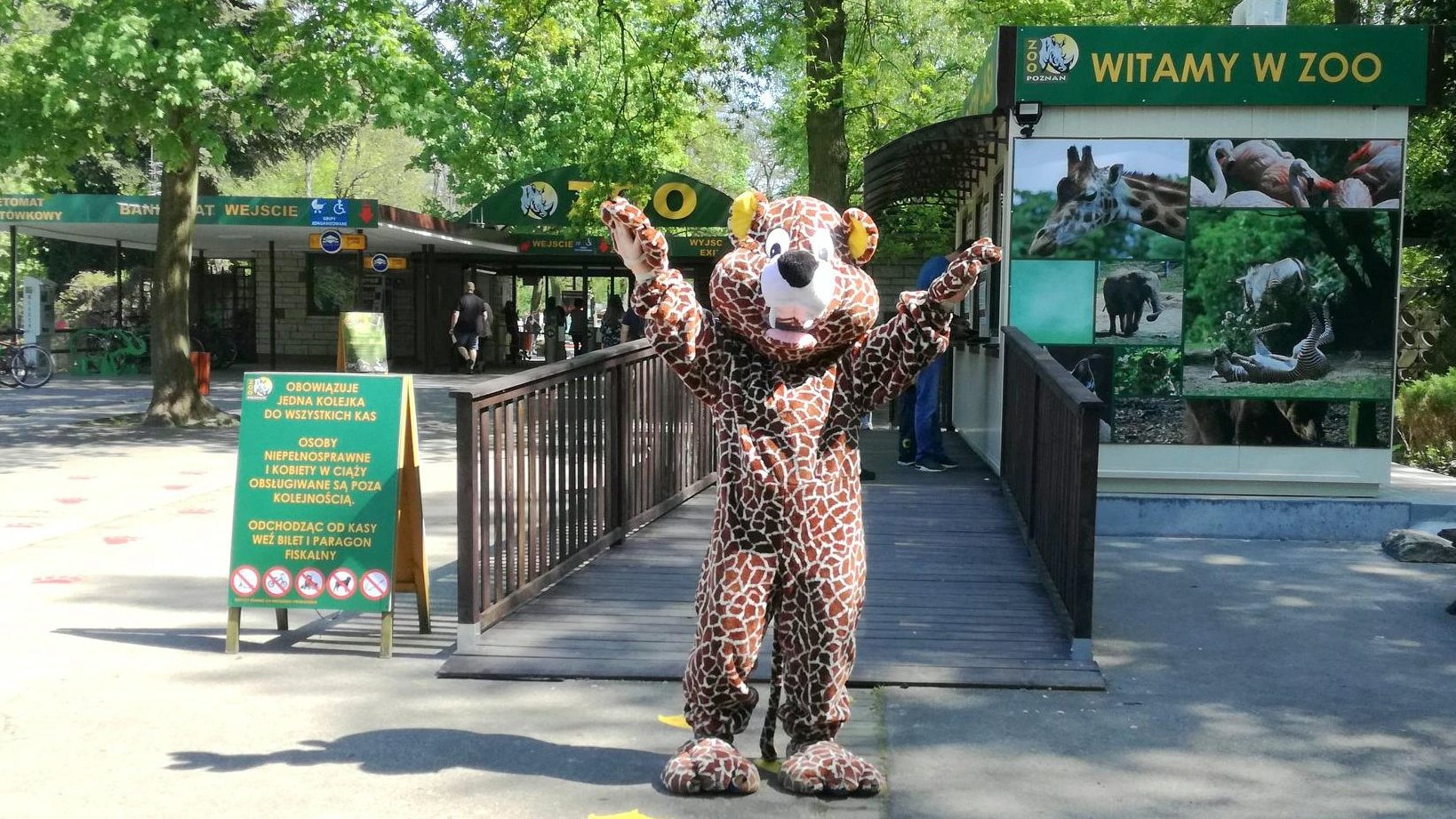 Zdjęcie przedstawia wejście do zoo. Na pierwszym planie widać osobę przebraną za geparda - stoi ona z otwartymi ramionami, w geście przywitania. W tle widać kasy, bramki wejściowe i napis "witamy w zoo". - grafika rozmowy