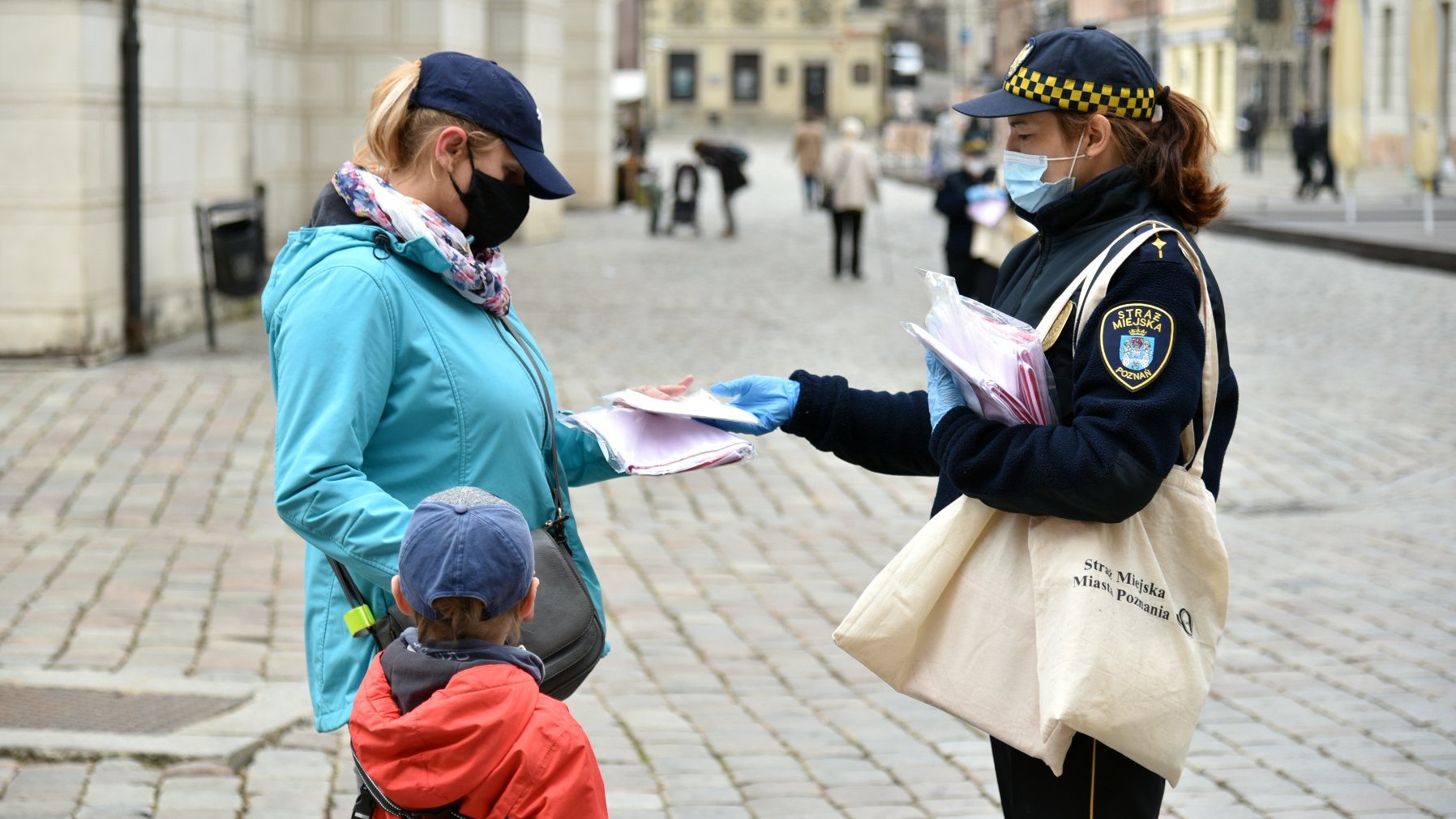 Galeria zdjęć przedstawia strażniczki miejskie rozdające mieszkańcom biało-czerwone flagi.