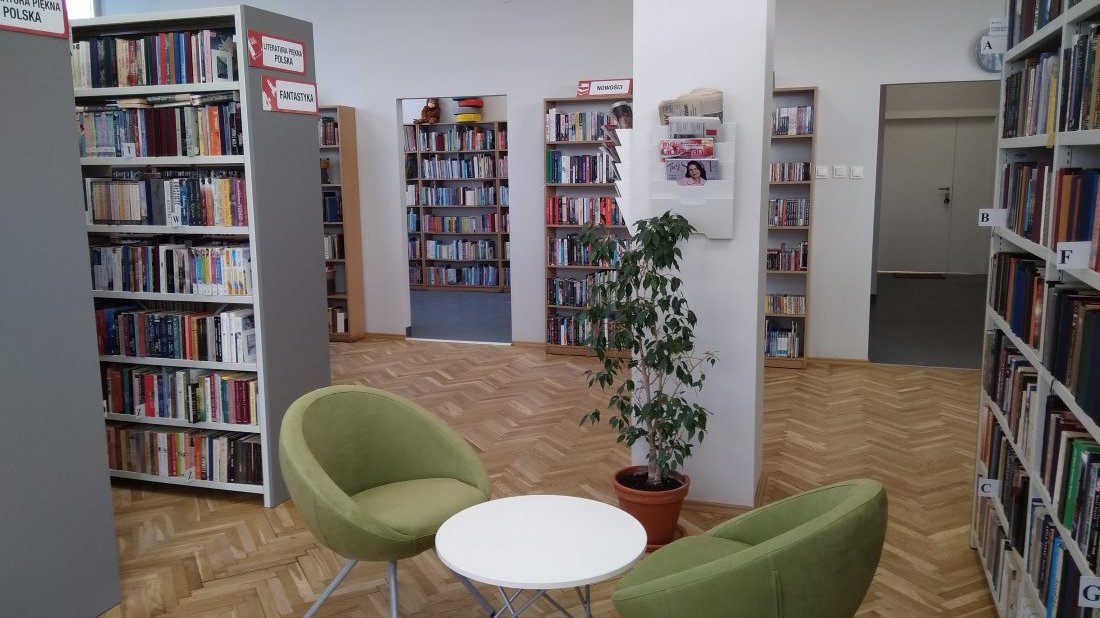 Zdjęcie przedstawia wnętrze biblioteki. Widać na nim półki z książkami oraz mały biały stoliczek z dwoma zielonymi krzesłami.