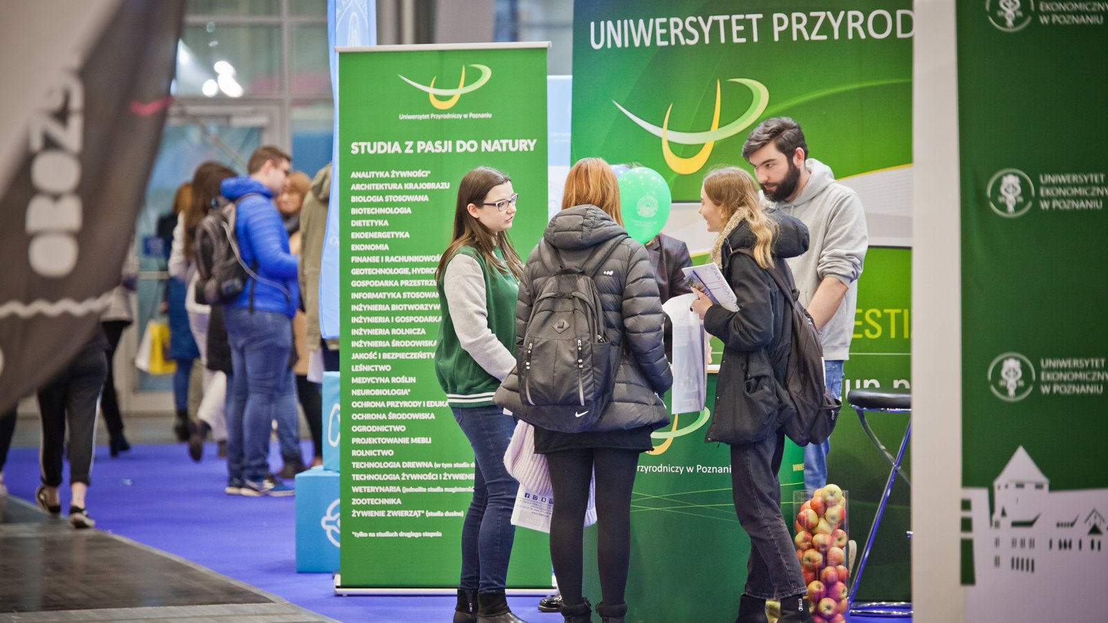 Zdjęcie przedstawia grupkę młodych ludzi stojących przed dużym zielonym banerem Uniwersytetu Przyrodniczego.