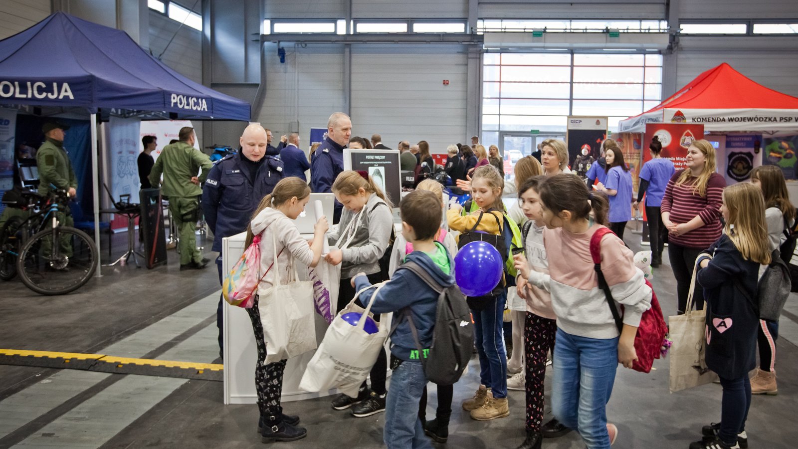 Zdjęcie przedstawia grupkę dzieci stojących przed stoiskiem policji. Dzieci mają ze sobą torby i balony. W tle widać innych ludzi.