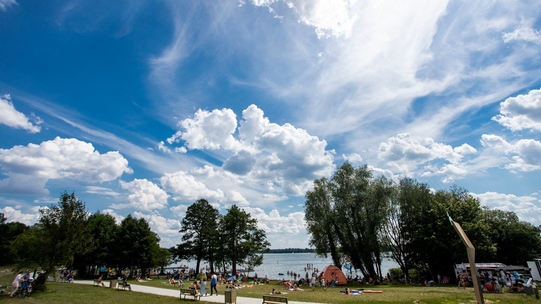 Zdjęcie przedstawia plażę wypełnioną ludźmi. Dalej widać też jezioro.