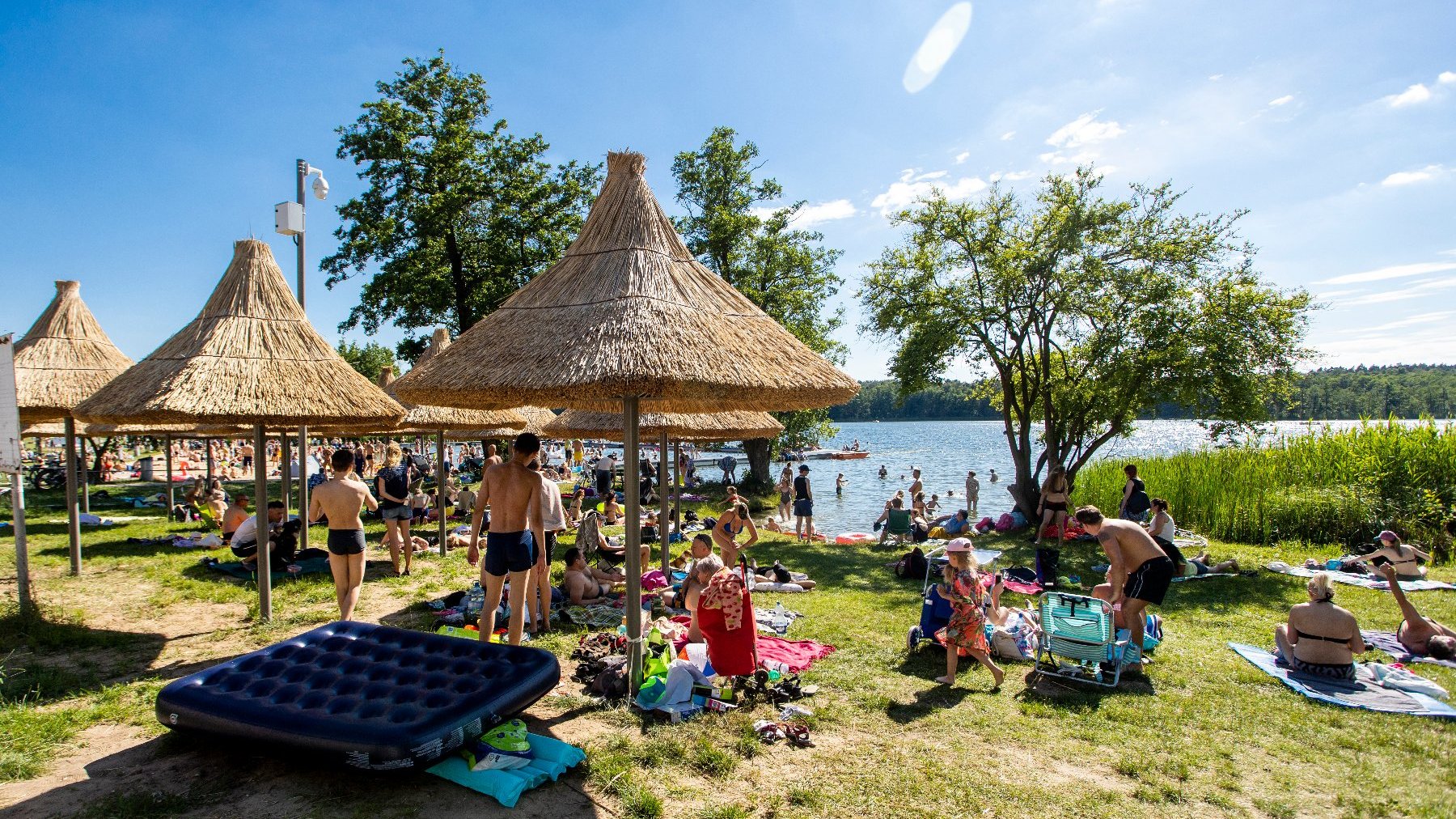 Zdjęcie przedstawia ludzi na plaży. Wielu z nich wypoczywa pod parasolami ze słomy. W oddali widać jezioro.