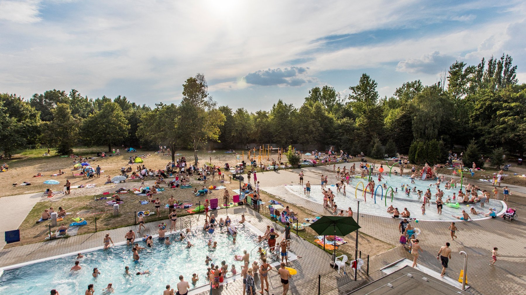 Ludzie wypoczywają na pływalni zewnętrznej. Niektórzy z nich kąpią się w jednym z dwóch basenów, inni leżą na plaży.