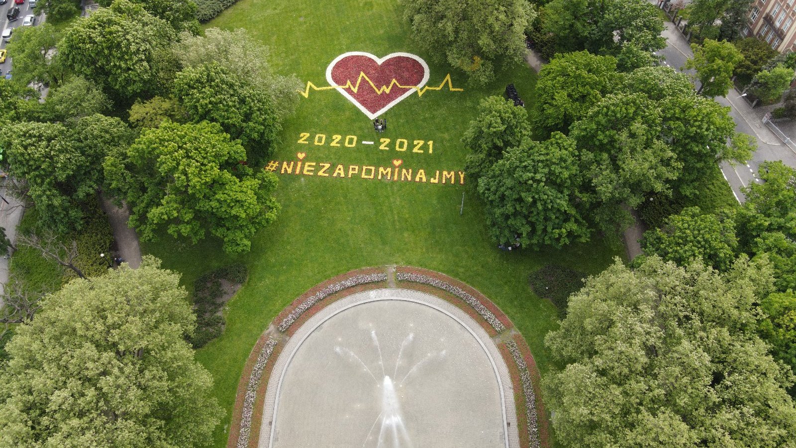 Zdjęcie przedstawia ułożone z kwiatów serce i napis "Nie zapominajmy". Wkoło widać drzewa, obok znajduje się fontanna. Fotografia została zrobiona z góry, za pomocą drona.