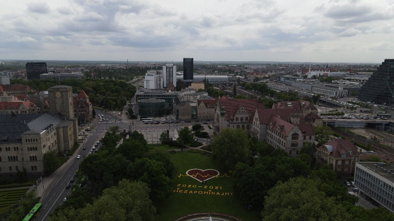 Zdjęcie przedstawia panoramę miasta widzianą z góry. Pomiędzy budynkami znajduje się park a tam ułożone z kwiatów serce i napis "Nie zapominajmy".