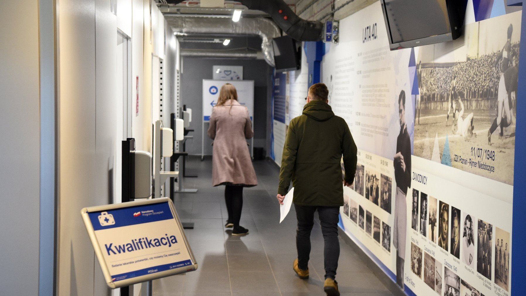 Na zdjęciu wnętrze punktu szczepień: korytarz, obok pomieszczenia do kwalifikacji, w korytarzu dwoje ludzi, widzianych z tyłu