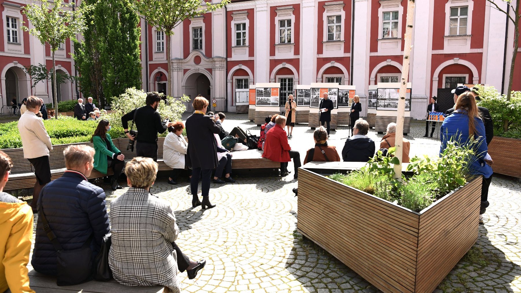 Dziedziniec urzędu miasta. Kilkanaście osób siedzi na ławkach, obserwują rozdanie nagród w konkursie. W tel budynek urzędu.