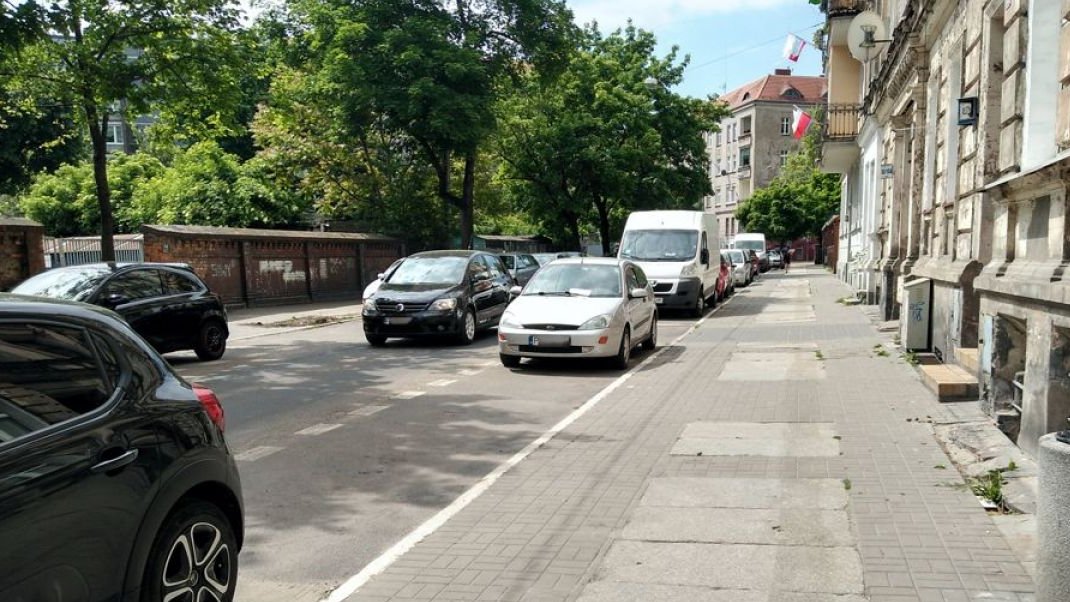 Zdjęcie przedstawia jezdnię i chodnik. Na poboczu drogi stoją zaparkowane samochody.