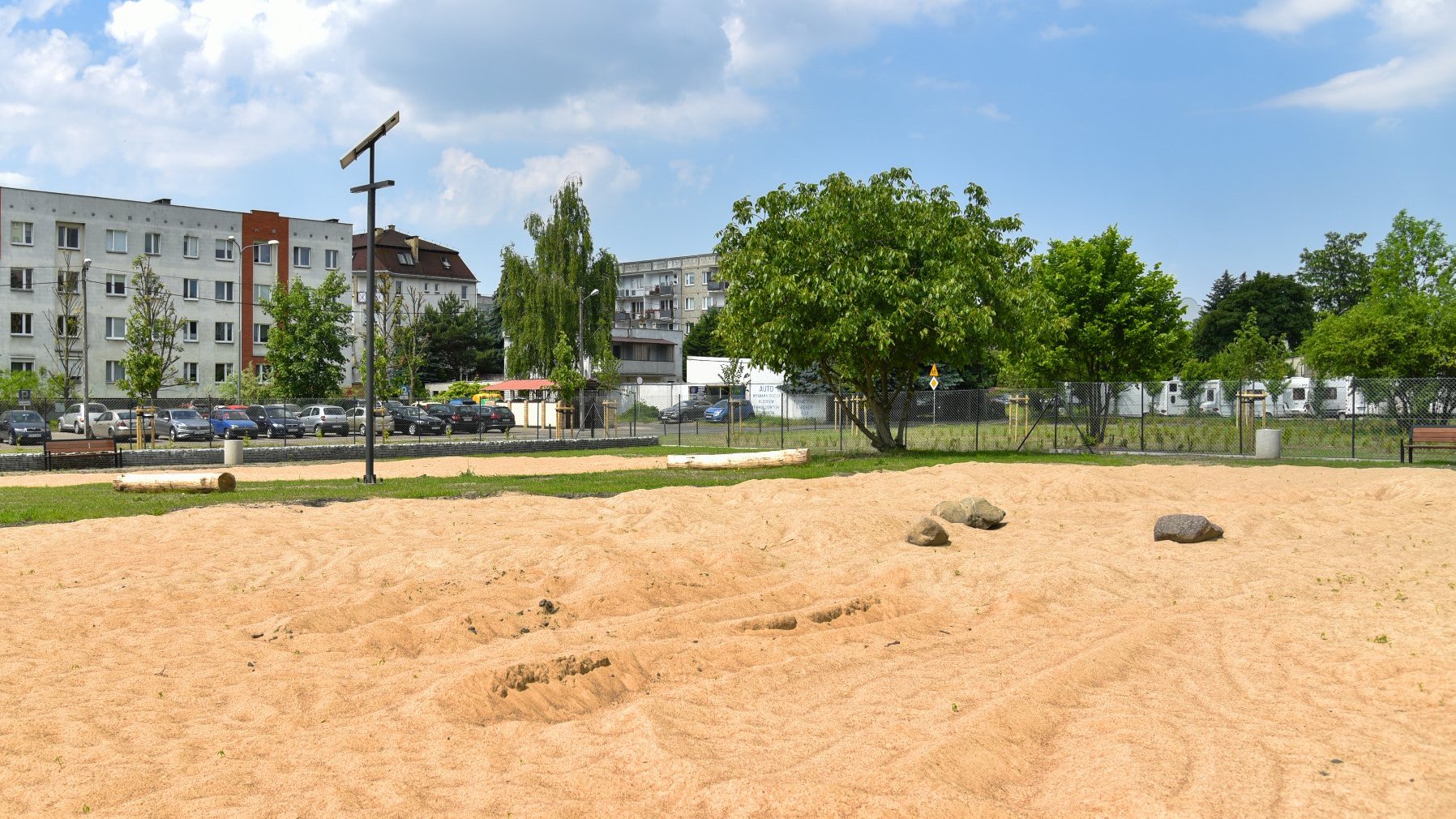 Zdjęcie przedstawia wybieg dla psów. Widać na nim m.in. piasek i kamienie. W tle znajdują się bloki i parking.