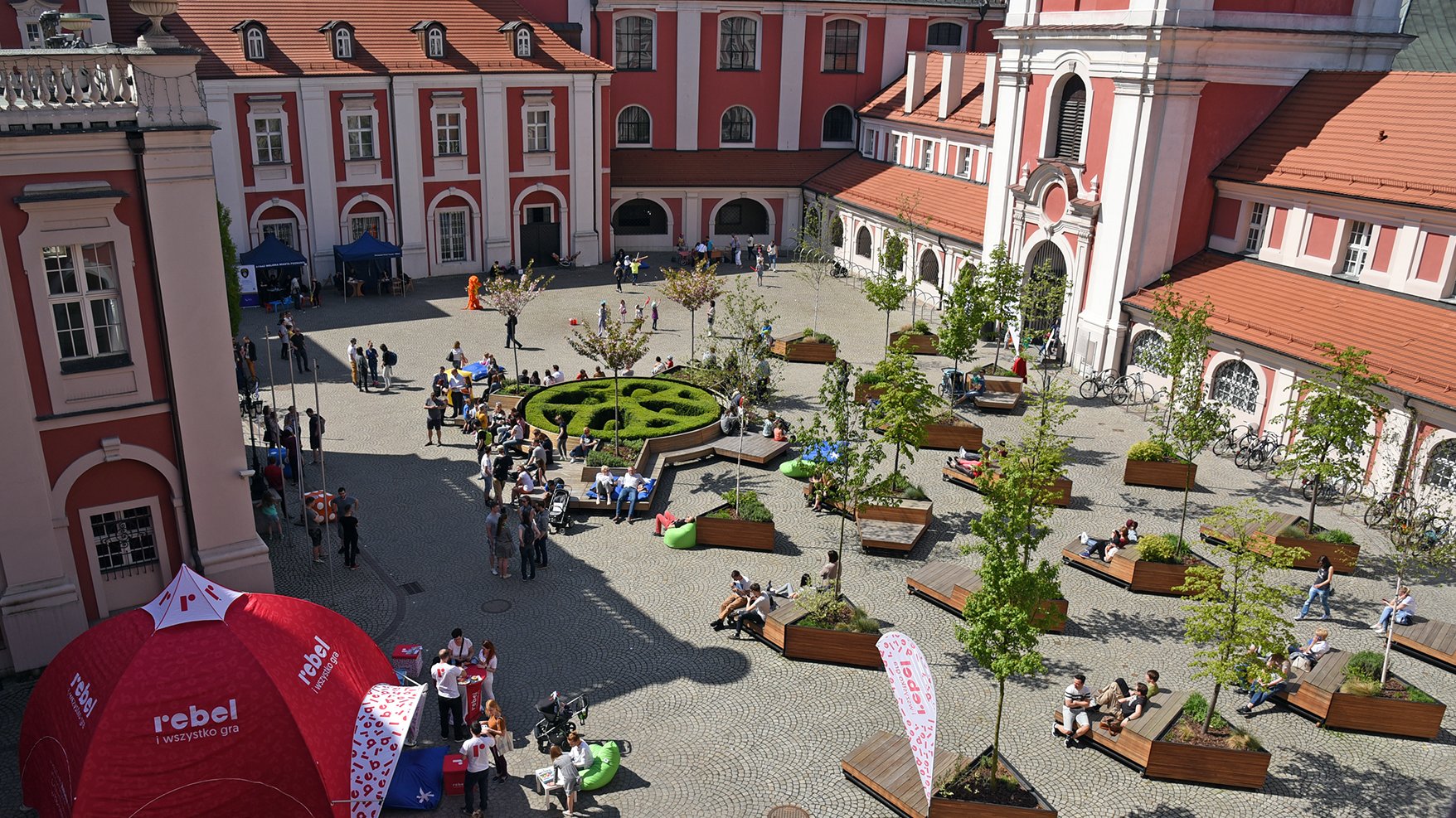 Zdjęcie przedstawia dziedziniec urzędu miasta. Na palcu widać sporo osób siedzących na ławkach i spacerujących. - grafika rozmowy