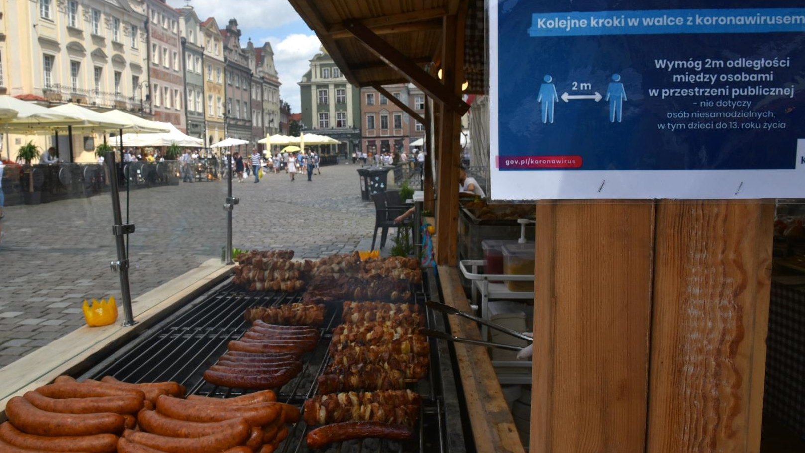 Zdjęcie przedstawia drewniany domek, w którym sprzedawane są szaszłyki i kiełbasy z grilla. W tle widać ludzi chodzących po Starym Rynku.